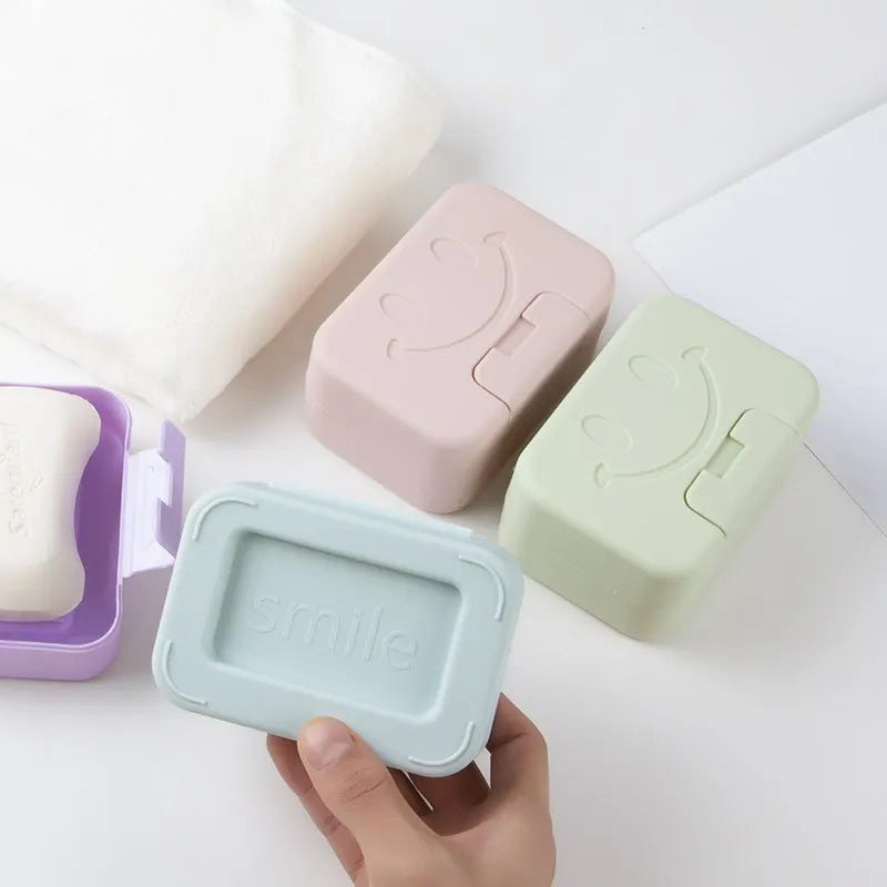 A hand holds a light blue smiley face lidded soap dish next to pink and green versions on a white surface.