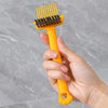 A hand holds an orange dual-sided hairbrush cleaner with rake teeth against a gray background.