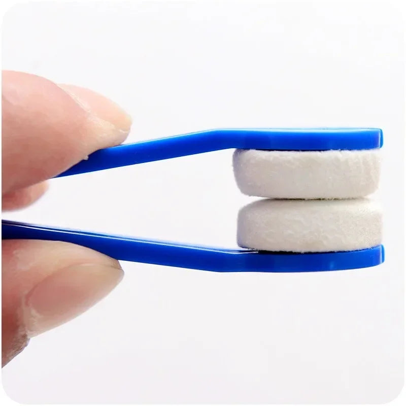 A hand holds a blue cleaning brush with two white microfiber pads for cleaning glasses.