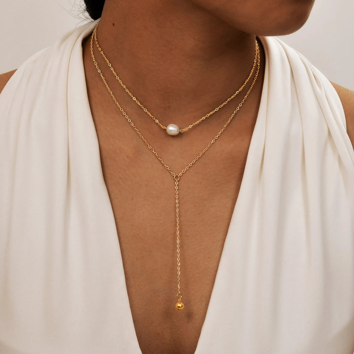 Una mujer vistiendo un collar lariat dorado superpuesto con una perla y un pequeño adorno dorado contra un vestido color crem