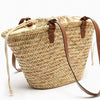 A beige handwoven straw tote bag with brown leather straps is shown against a white background.