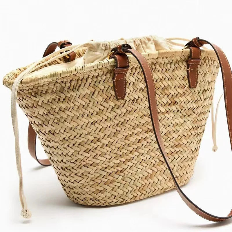 A beige handwoven straw tote bag with brown leather straps is shown against a white background.
