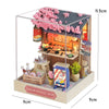 A miniature wooden dollhouse shop display featuring pink flowers and a counter, set within a clear acrylic box.