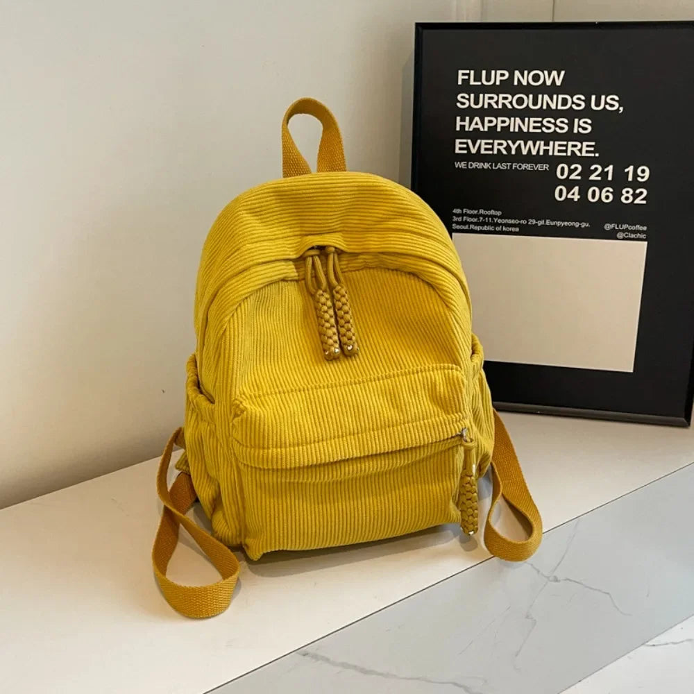 A yellow corduroy casual backpack is displayed on a white surface next to a framed sign.