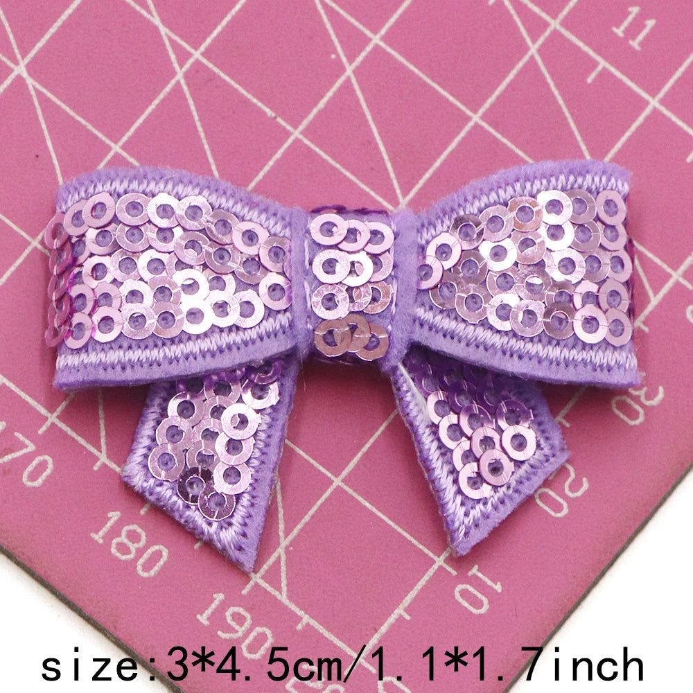Purple sequin bow patches for DIY crafts displayed on a pink grid background.