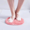 A pink silicone shower mat with exfoliating bristles is placed on a white surface, showing bare feet.