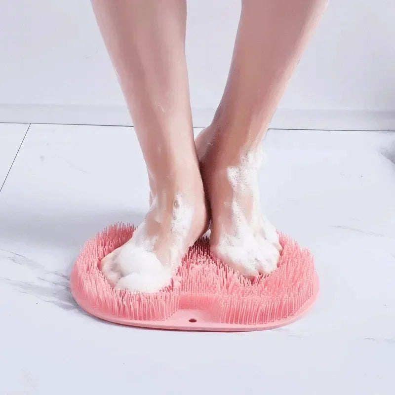 A pink silicone shower mat with exfoliating bristles is placed on a white surface, showing bare feet.