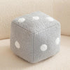 A gray, speckled dice-shaped plush pillow with white polka dots resting on a light beige surface.
