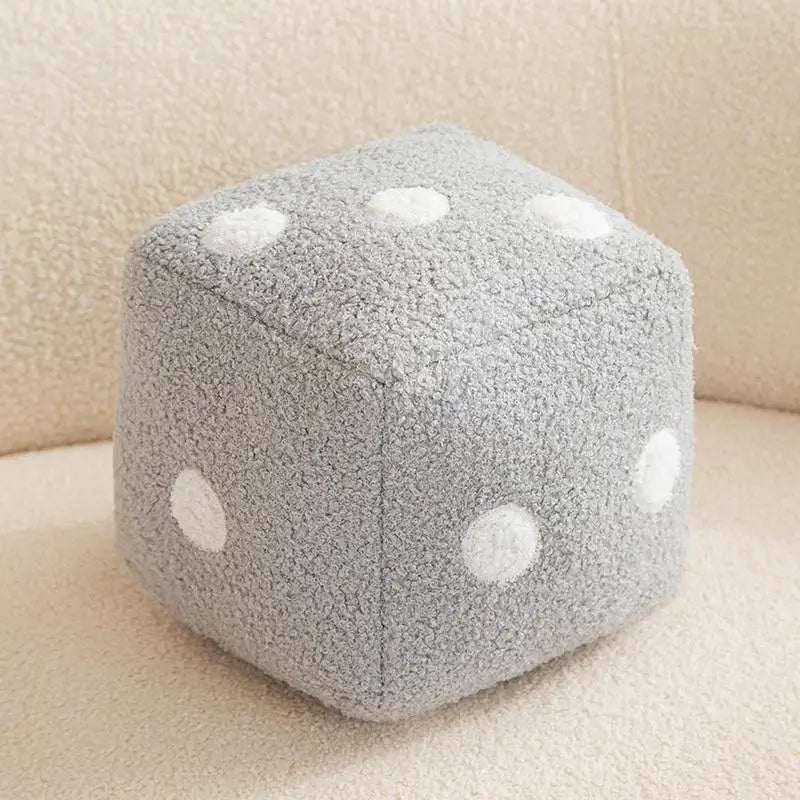 A gray, speckled dice-shaped plush pillow with white polka dots resting on a light beige surface.