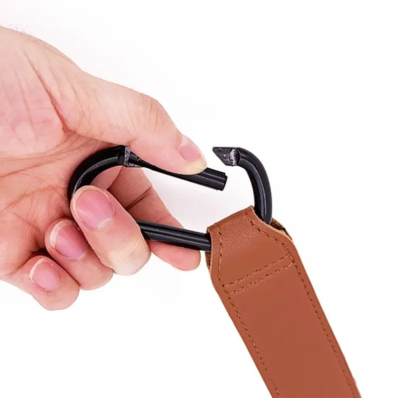 A hand is holding a black rotating stroller hook attached to a brown PU leather baby bag organizer.