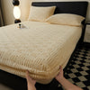 A close-up of a beige plush mattress cover with an elastic edge being held, resting on a black bed.