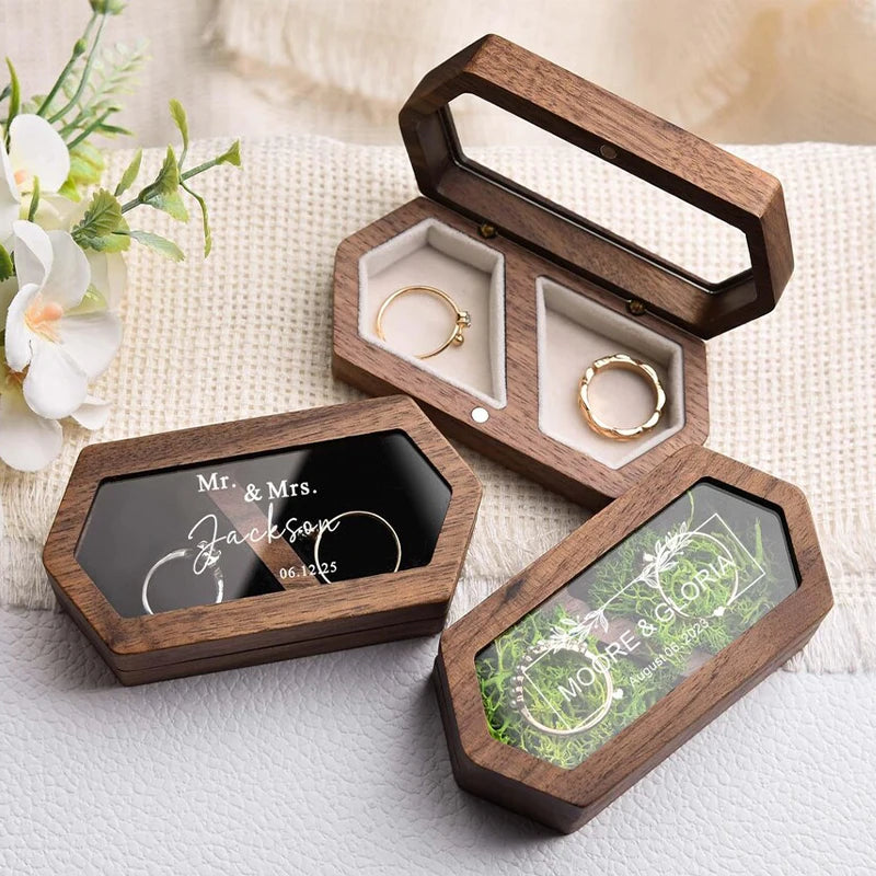 A wooden ring box with a lid open displaying gold rings and an engraved plaque on a white background.