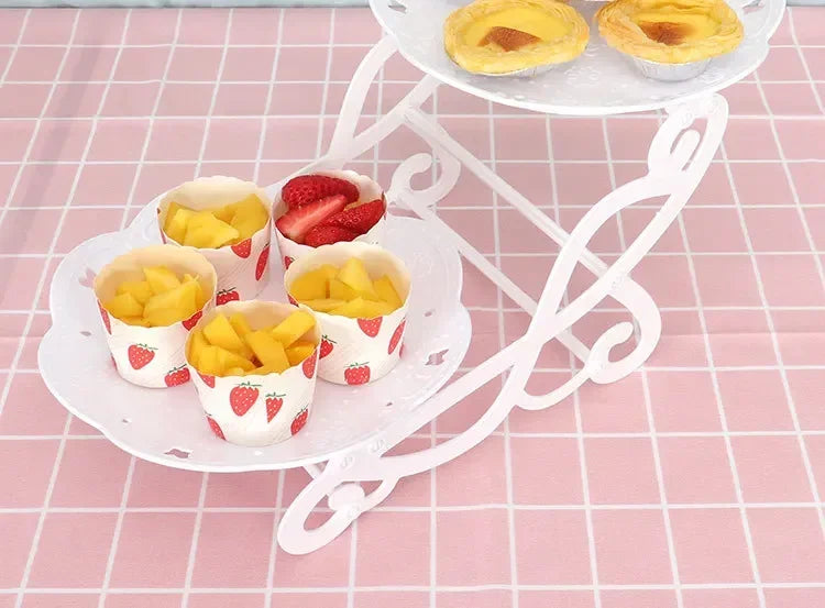 Ein weißer Plastik-Dessertständer hält mehrere kleine Tassen mit gelbem Obst und Erdbeerm desserts auf einem rosa karierten T