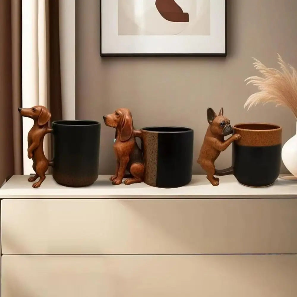 Three dachshund figurines with puppy handles rest on black and tan coffee mugs on a light shelf.