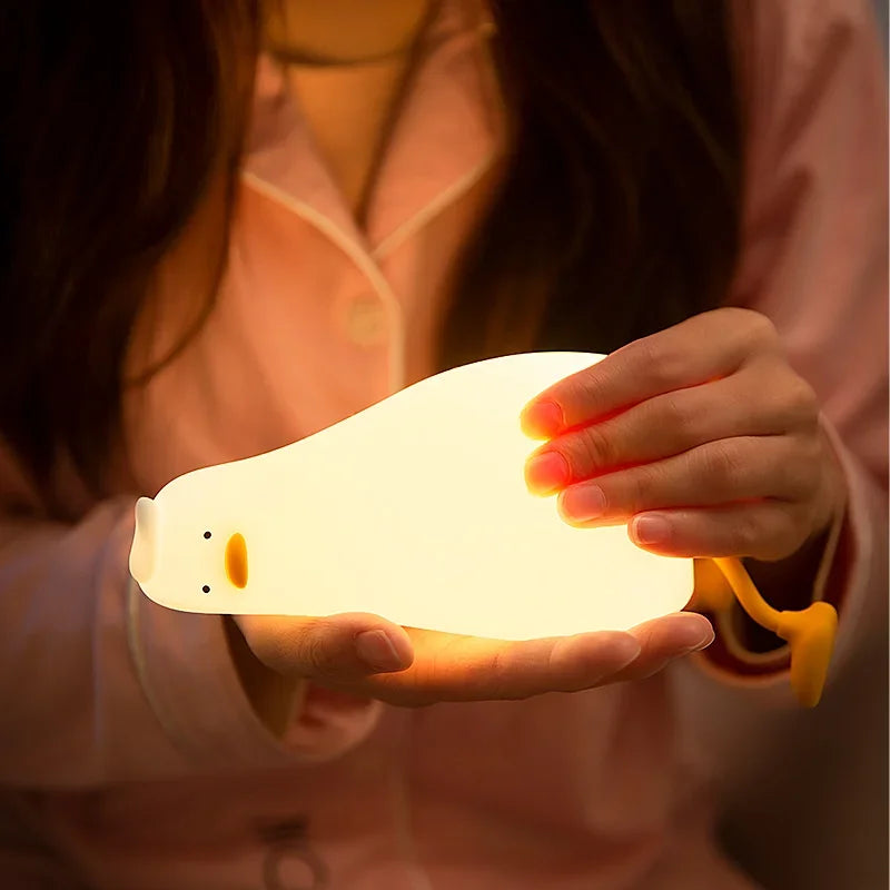 A hand holds a glowing white silicone duck touch night light against a soft pink background.