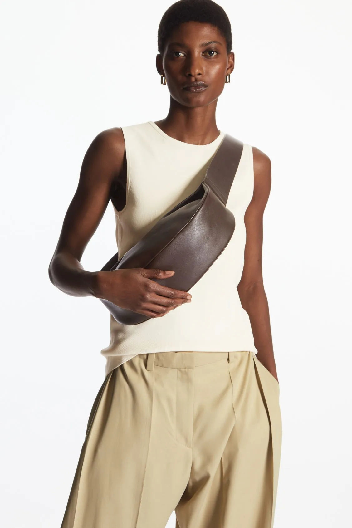 Model in cream top and beige trousers holding a brown leather crossbody bag