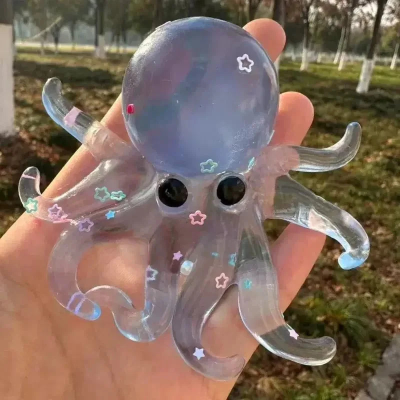 A clear silicone octopus squishy toy with purple and pink accents is held in a hand against a grassy outdoor background.