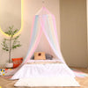 A white bed decorated with a rainbow hanging dome canopy mosquito net and pillows in a light room.