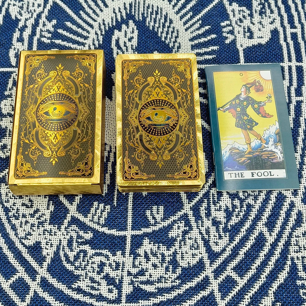 A set of gold foil tarot cards and guidebook featuring The Fool card on a blue patterned cloth.