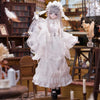 A delicate white lace dress doll with long silver hair and a feathered headdress displayed in a detailed miniature setting.