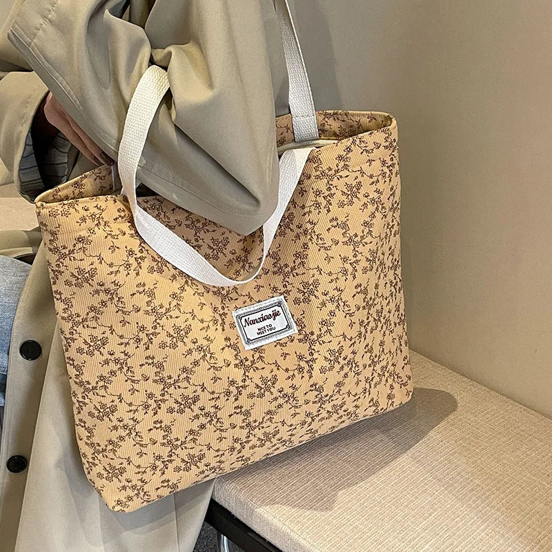 A beige corduroy tote bag with a floral pattern is held by a person in a light-colored coat.