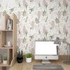 A computer monitor sits on a wooden desk in front of a wall covered with a boho leaf peel-and-stick wallpaper.