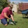 A man in a pink shirt is kneeling on grass, using a stainless steel weeder set to remove weeds.