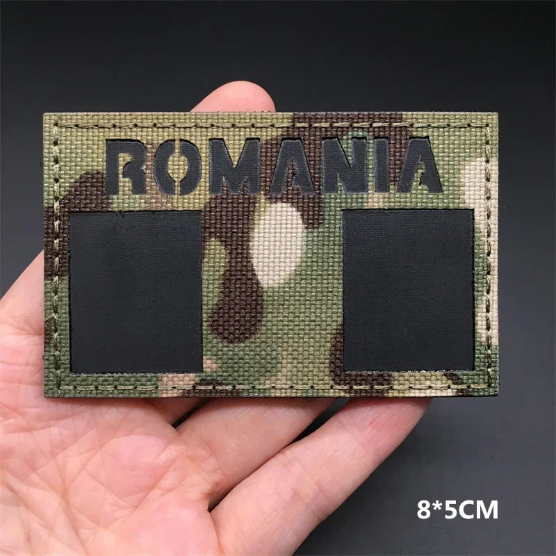 A rectangular camouflage patch with the word ROMANIA on a green and brown pattern is held in a hand.