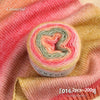 A roll of colorful gradient cake yarn showing pink, orange, and green strands on a pink fabric background.