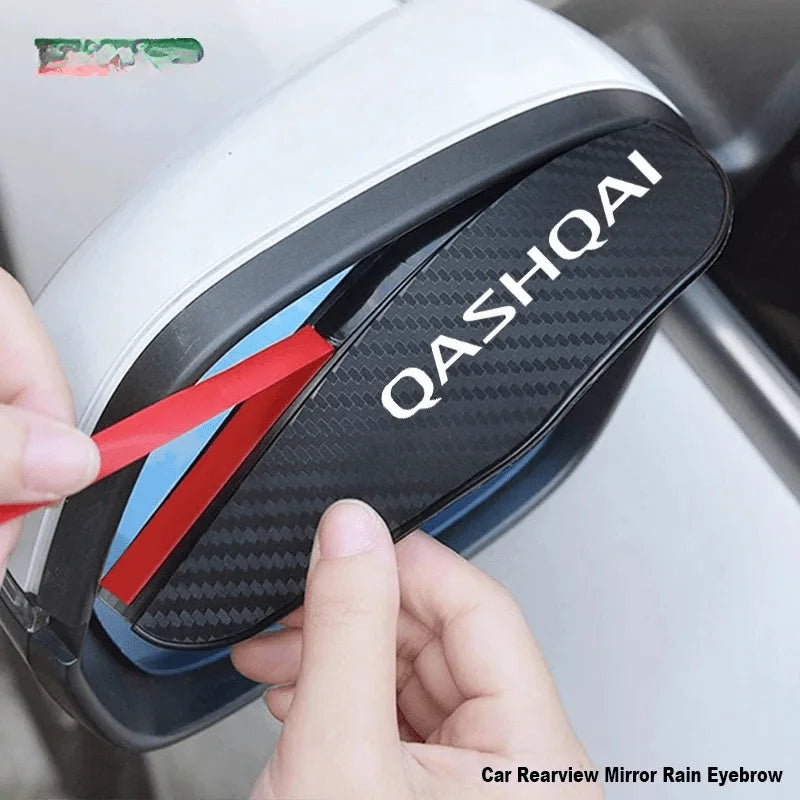 A hand is holding a black carbon fiber rain eyebrow guard for a Nissan Qashqai rearview mirror.
