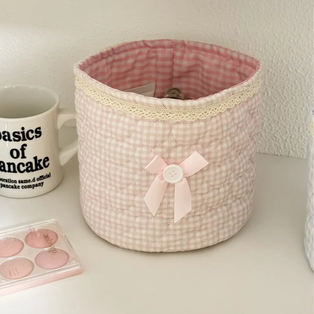 Cesta de almacenamiento acolchada rosa con ribete de encaje y cinta rosa, junto a una taza blanca y artículos rosas.
