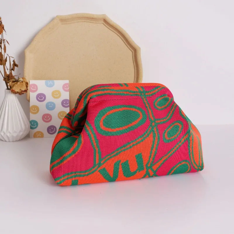 A hand-knit clutch bag featuring vibrant pink, orange, and green geometric patterns with pearl accents resting on a white sur