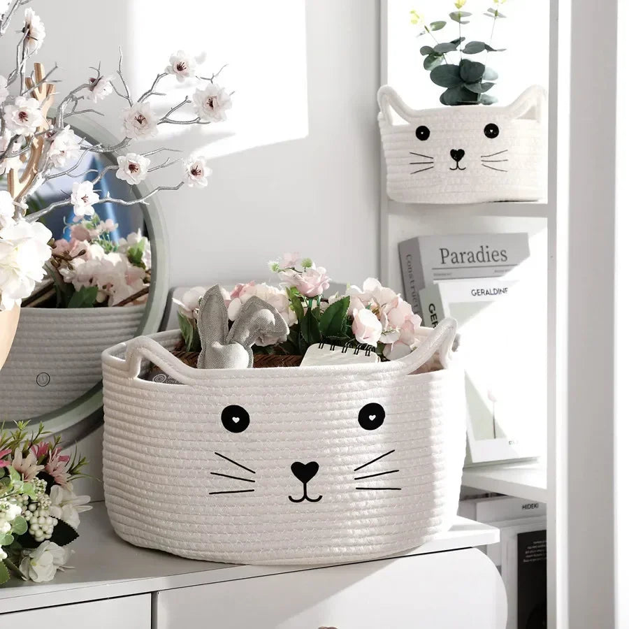 A white woven cotton rope storage basket shaped like a cat face with handles, holding pink and green flowers.