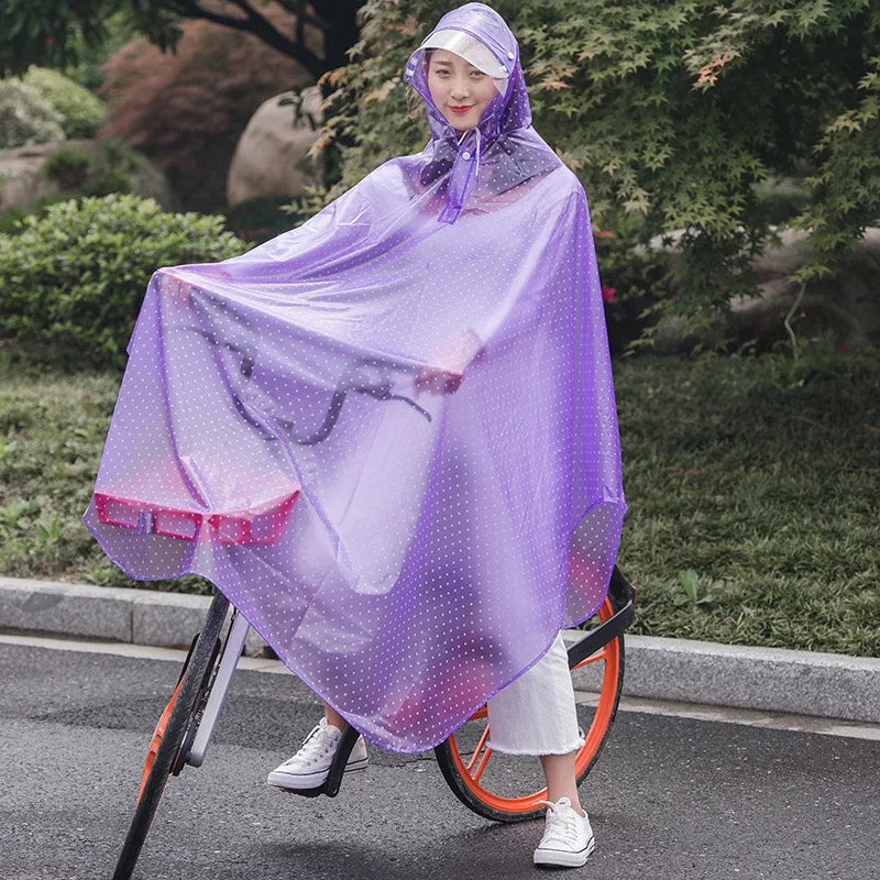 A woman wearing a purple transparent waterproof motorcycle rain poncho with a hood standing on a bicycle in a park setting.