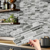 A hand holds a rectangular peel and stick waterproof tile sticker for kitchen or bathroom backsplash against a gray subway ti