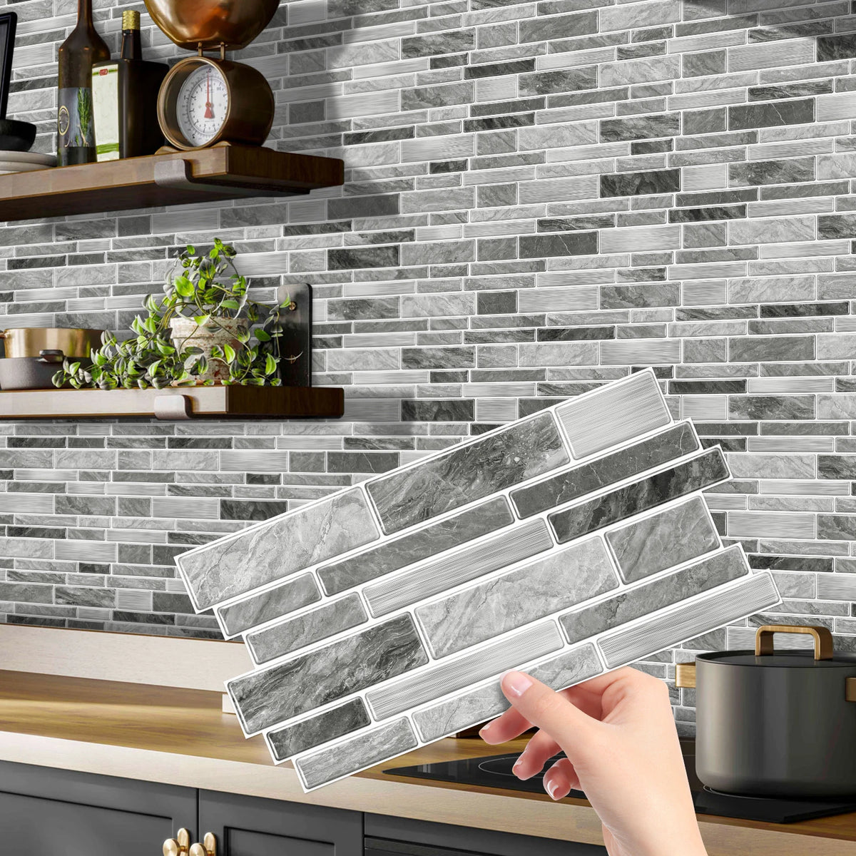 A hand holds a rectangular peel and stick waterproof tile sticker for kitchen or bathroom backsplash against a gray subway ti