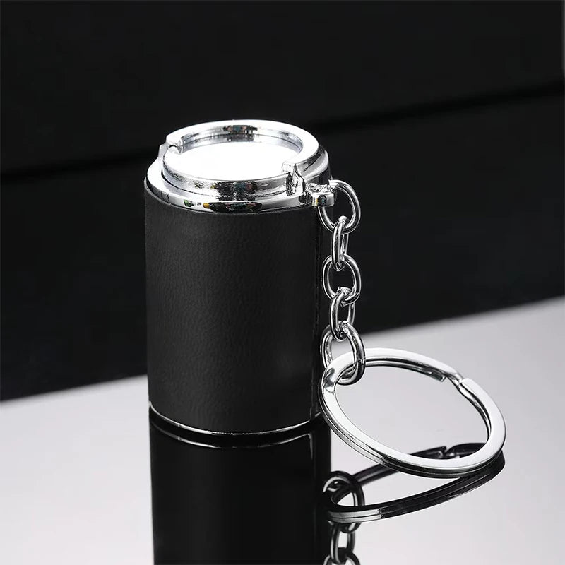 A black metal coin tube keychain with a silver chain and ring attached, shown on a reflective surface.