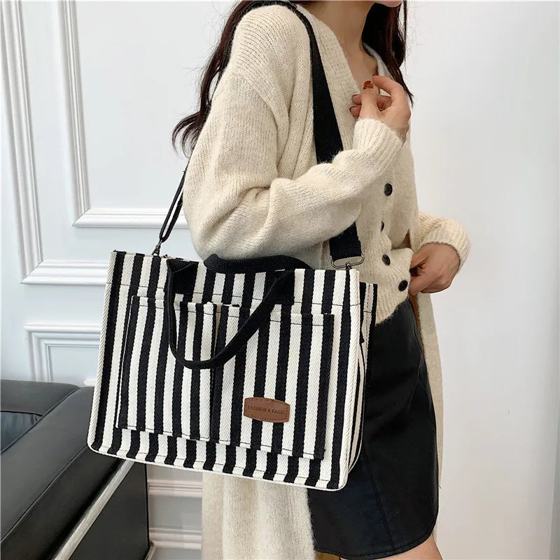 A woman holds a black and white striped canvas tote bag with inner pockets and a cream knit cardigan.