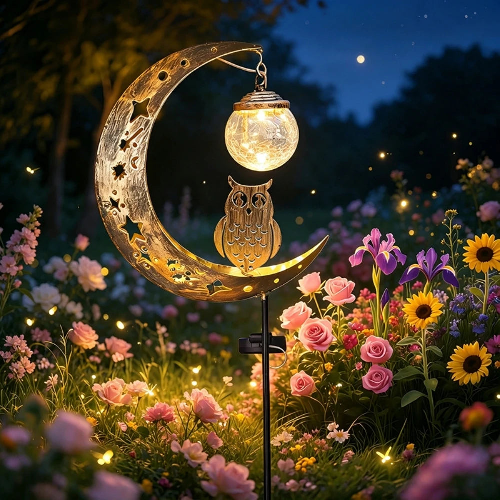 A solar owl garden light with moon decor hangs over a flower garden at twilight.