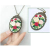 A silver-toned necklace featuring floral embroidery in pink, red, and cream tones on a circular pendant.