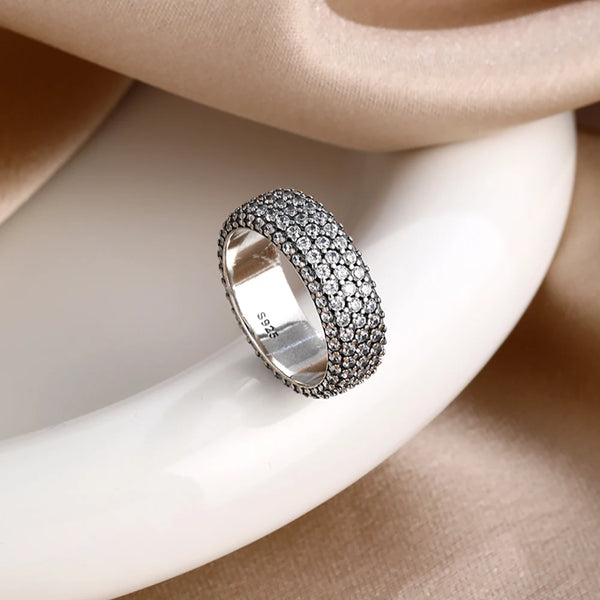 Nahaufnahme eines silbernen Rings, besetzt mit kleinen Diamanten, auf einer glatten weißen Oberfläche neben beigem Stoff