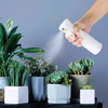 A hand sprays a white mist from a bottle onto green plants and foliage.