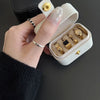 A hand holds an open cream-colored leather jewelry travel case containing several gold and silver rings.