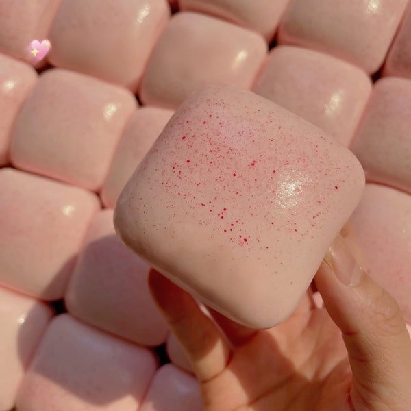 A hand holds a pink, square stress relief fidget toy with red glitter on it against a background of similar pink items.