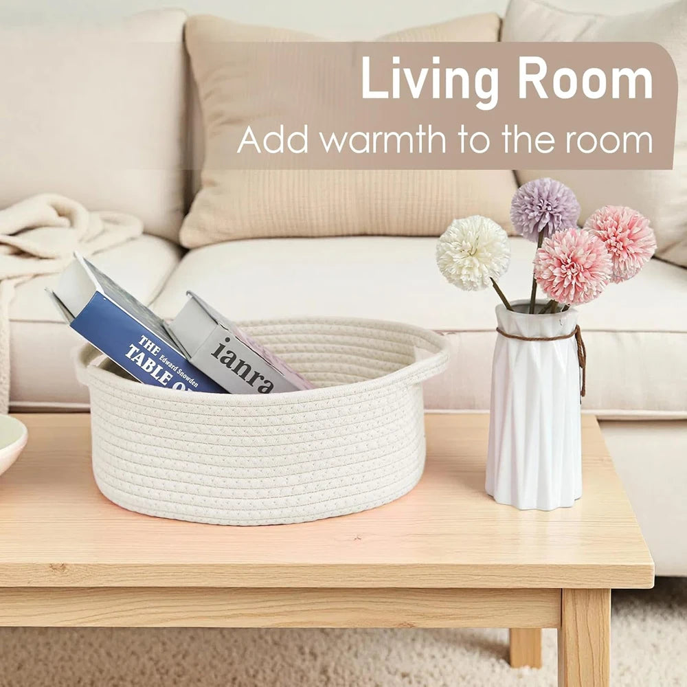 A white handwoven rope storage basket filled with magazines sits on a wooden table next to a pink floral arrangement.