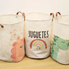 Three white fabric storage baskets with colorful cartoon prints of a unicorn, rainbow, and dinosaur on a wooden surface.