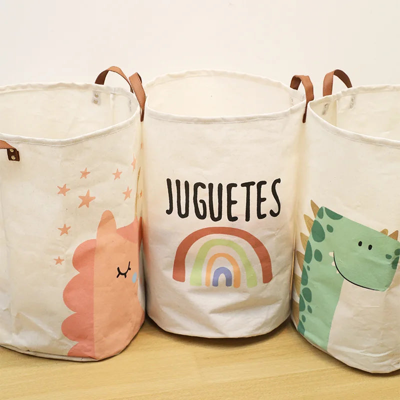 Three white fabric storage baskets with colorful cartoon prints of a unicorn, rainbow, and dinosaur on a wooden surface.