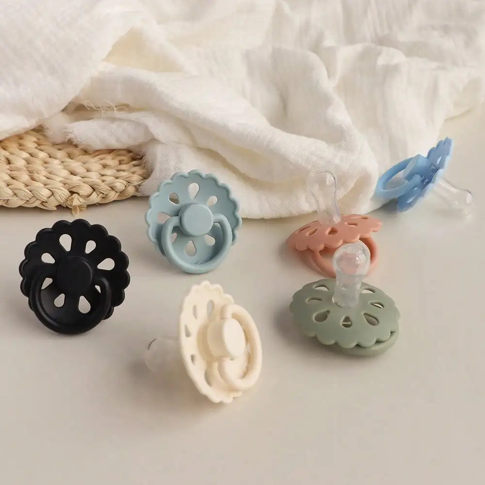 A collection of glow-in-the-dark silicone baby pacifiers in various pastel colors and shapes resting on a white cloth.