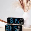A black LED digital wall clock displaying time, date, and temperature, next to a vase of dried grasses.
