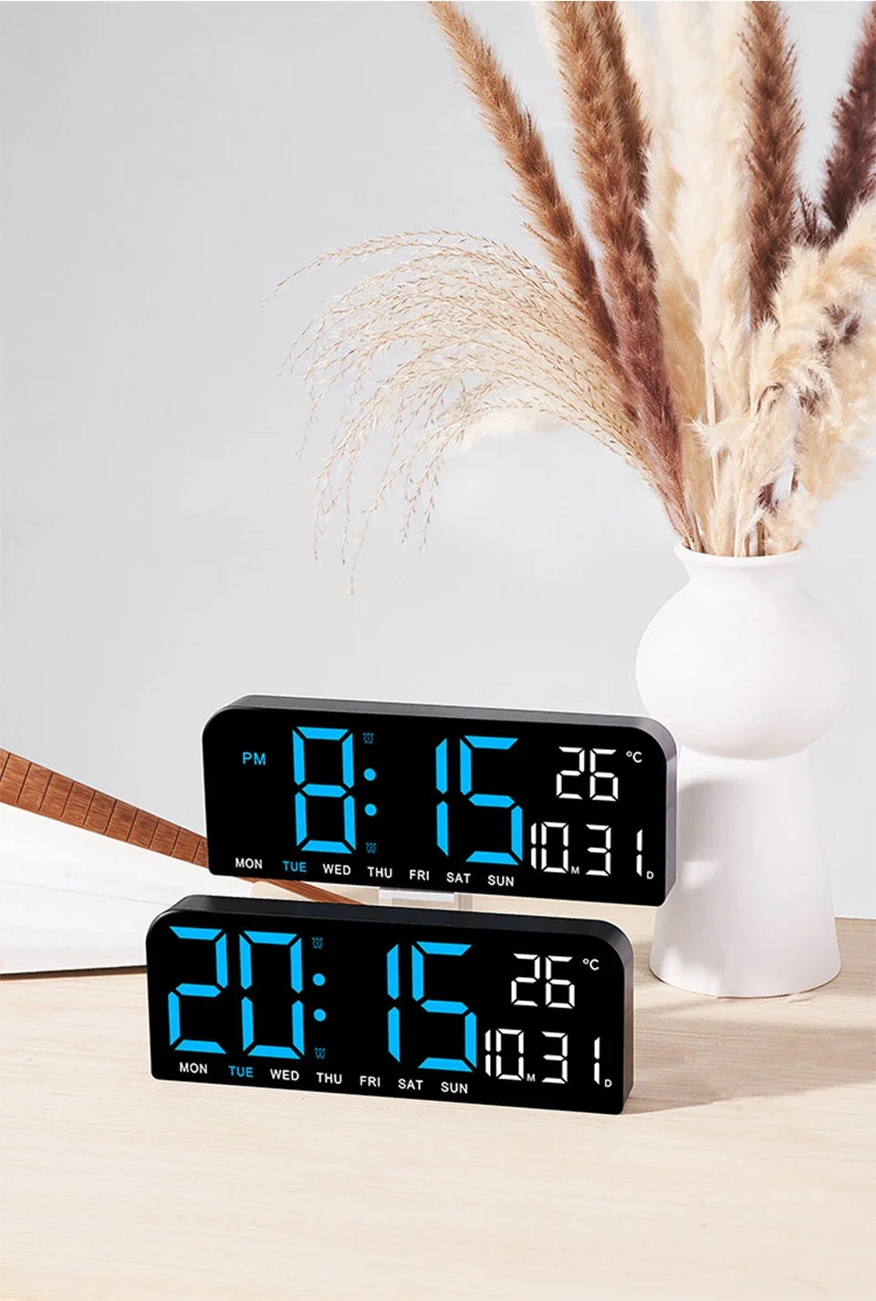 A black LED digital wall clock displaying time, date, and temperature, next to a vase of dried grasses.